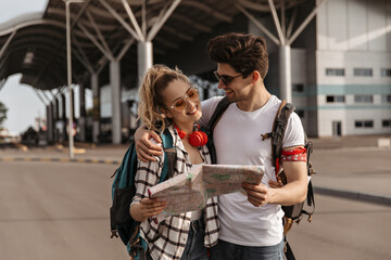 Fototapeta premium Cheerful woman and happy man in white tee-shirts smile and talk near airport. Travelers with backpacks hold map near airport.