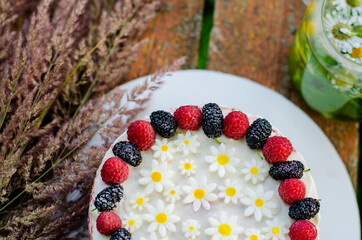 Cake decorated with berries and daisies. Teapot with chamomile and mint tea on a wooden table 