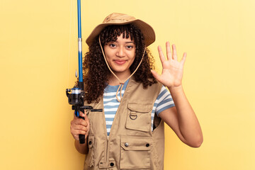 Young mixed race fisherwoman holding a rod isolated on yellow background smiling cheerful showing number five with fingers.