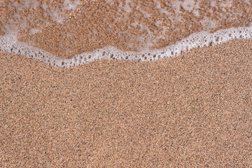 Background of a sandy beach with a sea wave. Foam from a wave on the sand. A foamy ocean wave on a seashell beach