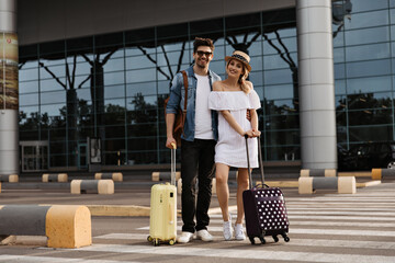 Blonde woman in hat, white dress and brunette man in denim jacket, sunglasses smiles and poses with suitcases near airport.