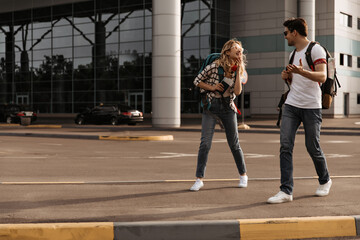 Charming woman in denim pants, plaid shirt and brunette man in jeans and white tee talks and smiles. Travelers poses with backpacks near airport.