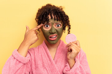 Young mixed race woman wearing a bathrobe holding a make-up remover sponge isolated on yellow background showing a disappointment gesture with forefinger.