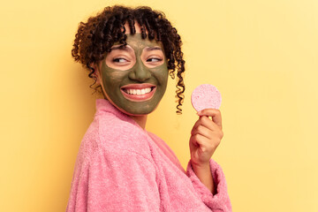 Young mixed race woman wearing a bathrobe holding a make-up remover sponge isolated on yellow background looks aside smiling, cheerful and pleasant.