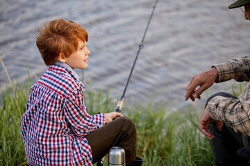 Grandfather and grandson fishing on river berth, senior man talking to kid boy. Redhead child in checekred shirt sit listening to talk on granddad, side view portrait