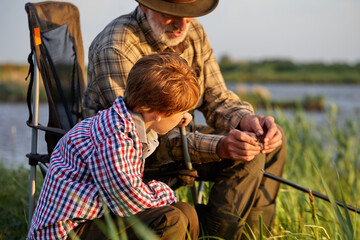 Senior man teach grandson how to put a worm on fishing hook, close-up, side view. Friendly family is preparing to fish together on lake, have talk. in countryside. copy space