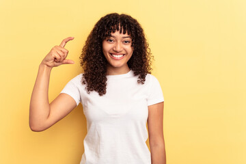 Young mixed race woman isolated on yellow background holding something little with forefingers, smiling and confident.