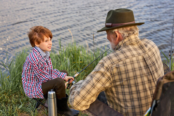 nice grandfather and grandson fishing together during camping trip by lake, copy space. side view on senior male and child having talk, adult man teach to fish, in nature, countryside