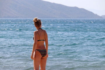 Slim woman in striped swimsuit standing in sea water and looking on mountain coast in mist, rear view. Beach vacation, perfect female body, summer leisure	