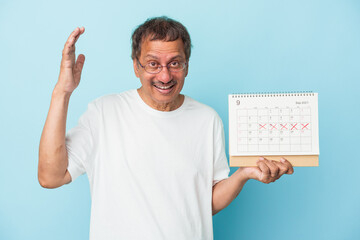 Senior indian man holding a calendar isolated on blue background receiving a pleasant surprise, excited and raising hands.