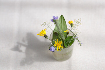 Wild flowers are arranged in a glass vase, with sunlight and shadows. A child's floral art with purple, yellow, blue and green colors on a beige background.