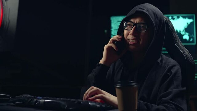 Asian Male Hacker Using Computer Hacking And Calling Phone
