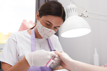 Professional manicurist wearing face mask, filing nails of client