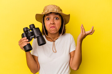Young mixed race explorer woman holding binoculars isolated on yellow background shrugs shoulders...