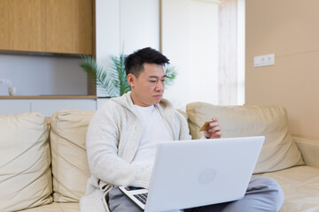 Fototapeta premium young happy asian man shopping online in internet store using laptop computer and credit card. male enters data of paying for services or tickets in application sitting on couch at home living room