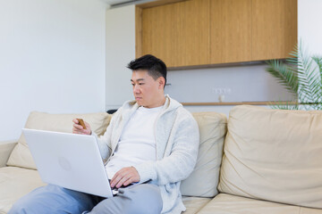 young happy asian man shopping online in internet store using laptop computer and credit card. male enters data of paying for services or tickets in application sitting on couch at home living room