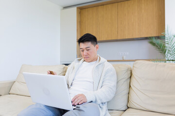 young happy asian man shopping online in internet store using laptop computer and credit card. male enters data of paying for services or tickets in application sitting on couch at home living room