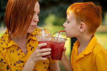 Red-haired young boy and mom are drinking red lemonade. Summer outdoor recreation.