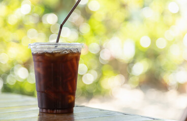 Closeup of take away plastic cup of iced black coffee Americano on wooden table with green nature background.