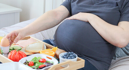 Obraz na płótnie Canvas Pregnant woman and breakfast on a wooden tray. A pregnant woman and breakfast on a wooden tray lie on the bed in the bedroom, close-up side view.