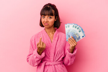 Young mixed race woman wearing a bathrobe holding bills isolated on pink background pointing with finger at you as if inviting come closer.