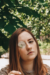Redhead girl with freckles holds a white dandelion seed head on a background of foliage on a sunny day. Young woman with white flower in her hands on the street