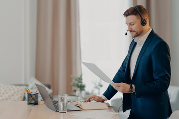 Serious man excutive worker gives presentation of business plan holds paper document stands near desktop dressed formally uses modern laptop and headset for online conference. Social distance working