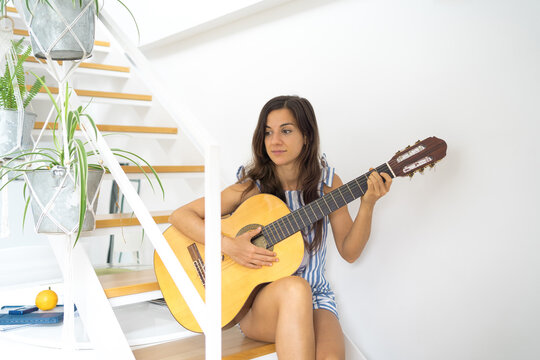 young woman sitting in modern apartment on stairs playing guitar
