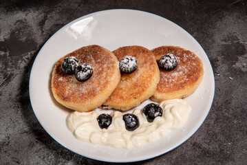 Cheesecakes (cottage cheese pancakes) with sour cream and blueberries on a white plate. Healthy tasty breakfast. Close-up