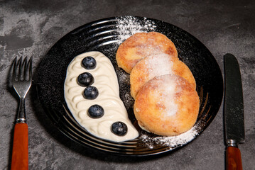 Cheesecakes (pancakes) with sour cream and blueberries on a black plate. Healthy tasty breakfast. Close-up