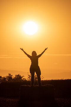 backlit photograph of the silhouette of a woman standing on a straw bale with her arms outstretched and the sun behind her