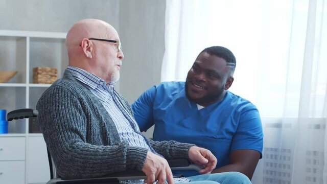 African-American caregiver and old disabled man in a wheelchair. Professional nurse and handicapped patient in a nursing home. Assistance, rehabilitation and health care.