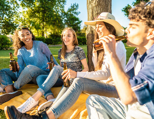 Happy friend group outdoor in city park drinking beer from bottle celebrating sitting on grass. Carefree young smiling people having fun in nature with alcohol at sunset enjoying a picnic in the grass