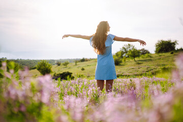 Naklejka na ściany i meble Young woman in stylish summer dress feeling free in the field with flowers in sunshine. Nature, vacation, relax and lifestyle. Summer landscape.