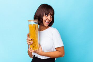 Young mixed race woman holding pasta jar isolated on blue background looks aside smiling, cheerful and pleasant.