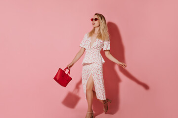 Fashionable blonde girl in red sunglassws, modern heels and light cool dress posing with trendy handbag on pink background..