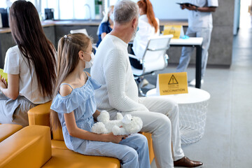 Child Girl Patient With Toy Sit In Hospital Waiting For Vaccination, Looking At Side, Scared And Frightened, Came With Grandfather, Side View. Copy Space