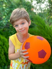 A cheerful, smiling little boy with a bright orange soccer ball against a background of green trees. Children's ball games in nature in the fresh air.