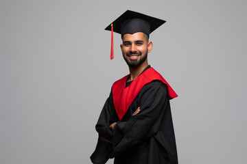 Smiling indian male graduate student in mortar board and bachelor gown with diploma over grey...