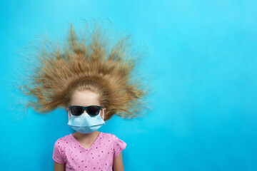 Beautiful European girl in a blue protective medical mask, sunglasses and a pink T-shirt lies with her blond hair loose in a semicircle on a uniform blue background, protection from covid, copy space
