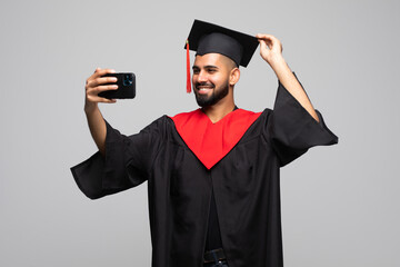 College indian graduate man taking a selfie with cell phone holding a diploma isolated on white...