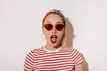 Shocked woman with red lips in sunglasses, cool earrings and striped modern t-shirt looking into camera on isolated backdrop..