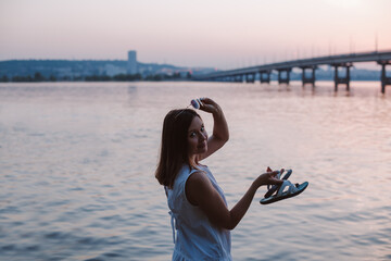 a slender brunette woman stands half-turned, takes off her sunglasses at sunset with a view of the city, the bridge and the river. 