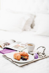 Breakfast in bed. Croissants with fresh berries, cup of coffee and flower on chic silver tray and blurry open magazine on white beautiful bed. Room service. 
