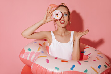 Obraz na płótnie Canvas Charming woman with trendy makeup on round earrings and white clothes posing with donut and swimming rings on pink backdrop..