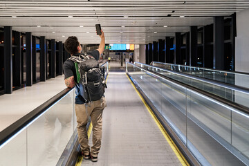 Obraz premium latin male tourist in mask advancing on airport treadmill with social distancing while making a video call during coronavirus pandemic or covid19 virus, concept of new normal