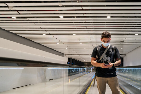 latin male tourist in mask advancing on airport treadmill with social distancing while making a video call during coronavirus pandemic or covid19 virus, concept of new normal