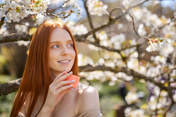 Beautiful sexy sensual woman with fresh health skin holding cream bottle in hands, in nature at spring, near blooming trees, sunny day. skincare, spa, natural cosmetic product concept
