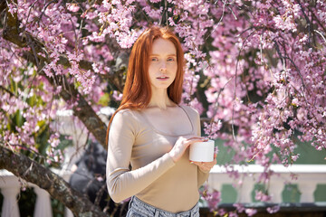 Close-up portrait of young beautiful redhead woman putting cream from cream bottle jar at spring park with blooming trees, sunny day. skincare, beauty, people lifestyle concept