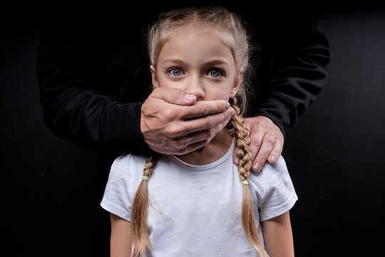 man kidnapping female child. The concept of kidnapping and child trafficking. Close up portrait of frightened child in white t-shirt looking at camera with wide opened eyes, isolated on black studio